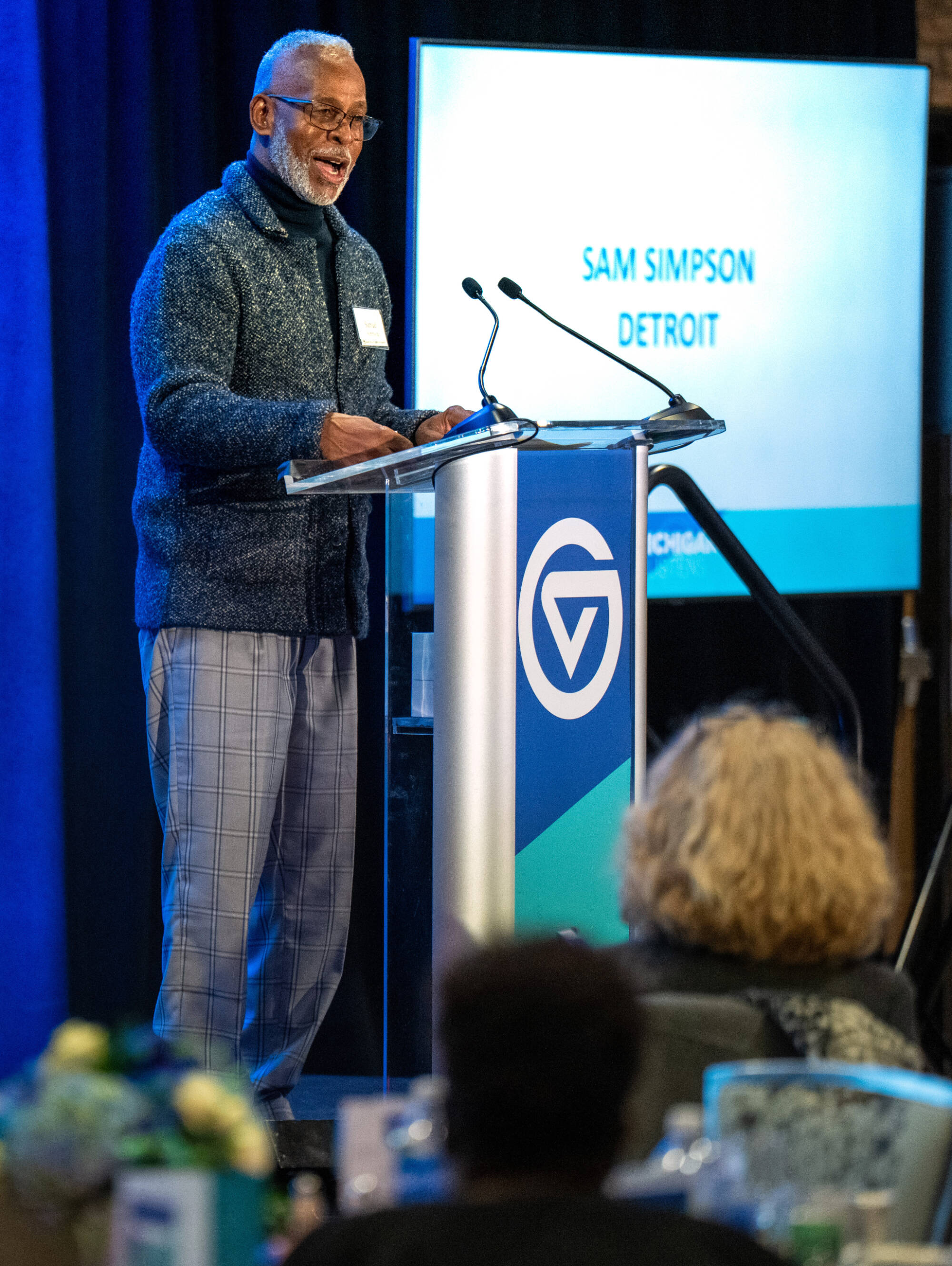 A man at the podium speaking with a slide with "Sam Simpson Detroit" in the background.